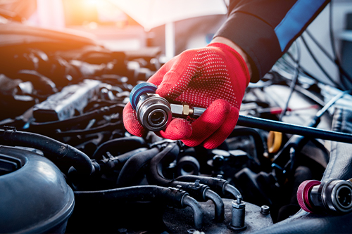 Car air condition repair in Los Angeles Ca at Sean's Auto Care. Image of mechanic hand wearing red protective glove holding A/C testing machine over car engine.