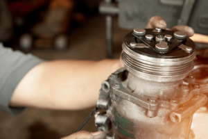 Auto air conditioner repair in Van Nuys, CA. Looking for an auto ac shop near you? Visit Sean's Auto Care. Image of mechanic holding the compressor of an auto ac system that came in the shop for repair.